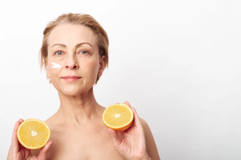 Mature woman holds two halves of an orange while applying cream on her face. Concept of natural skincare, healthy aging, vitamin care, and organic beauty treatment in bright studio setting.