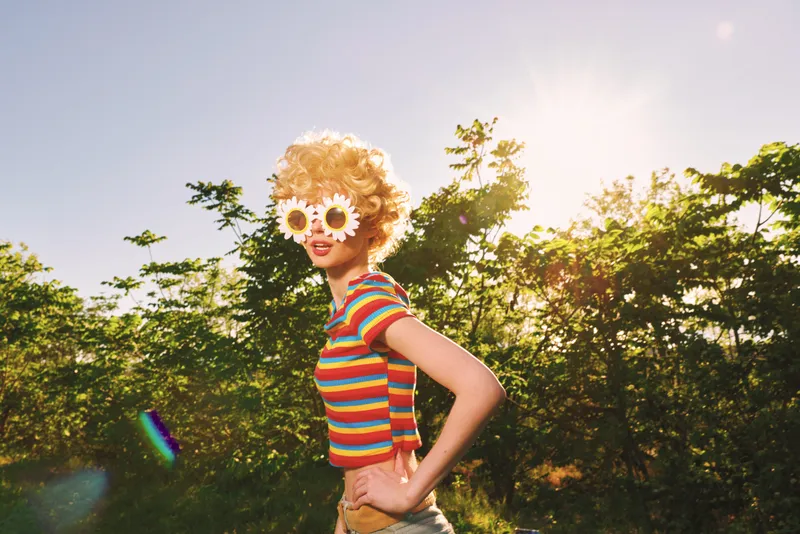 Cheerful woman outdoors, wearing striped shirt and flower sunglasses, enjoying warm sun in a lush nature setting with greenery and a soft breeze