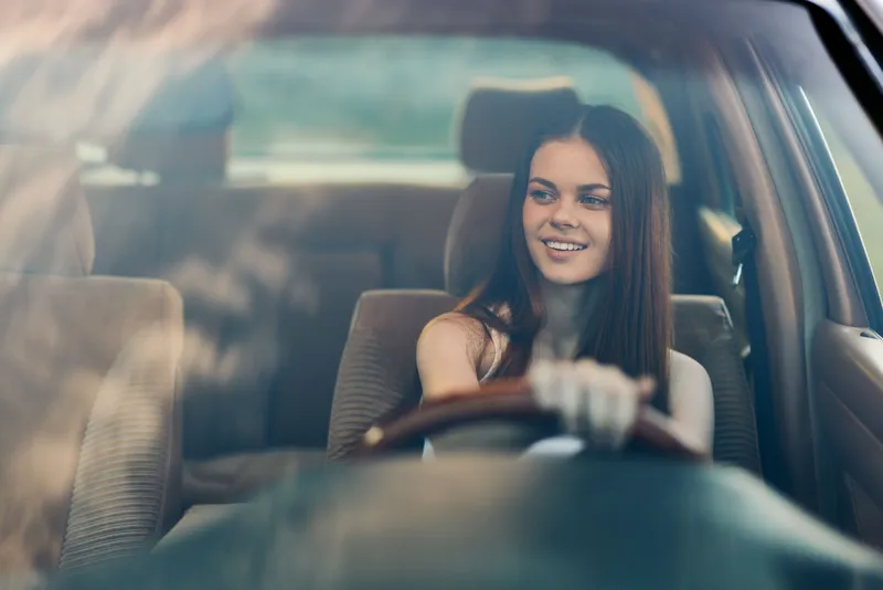 Enjoying a drive, a young woman smiles while sitting in a car, showcasing joy and freedom on a sunny day, ideal for concepts related to travel and adventure.