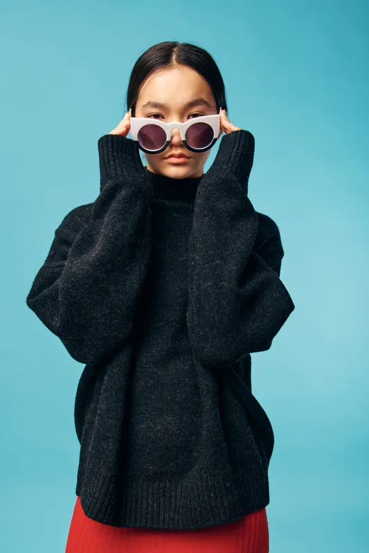Young woman wearing oversized black sweater and white round sunglasses posing against blue background. Stylish fashion portrait with casual winter clothing and confident expression.