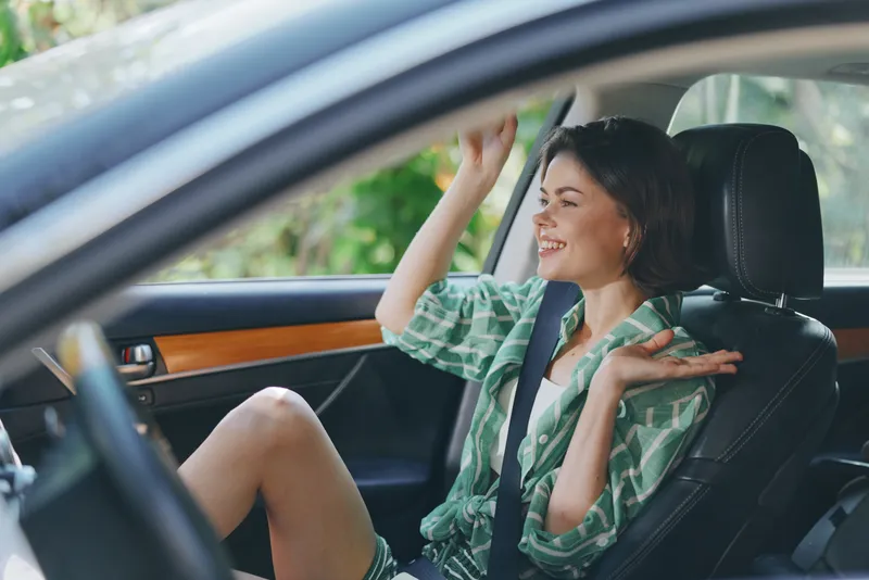 A woman sits inside a car with a relaxed smile, wearing a green shirt, seatbelt fastened, enjoying a sunny daytime ride.