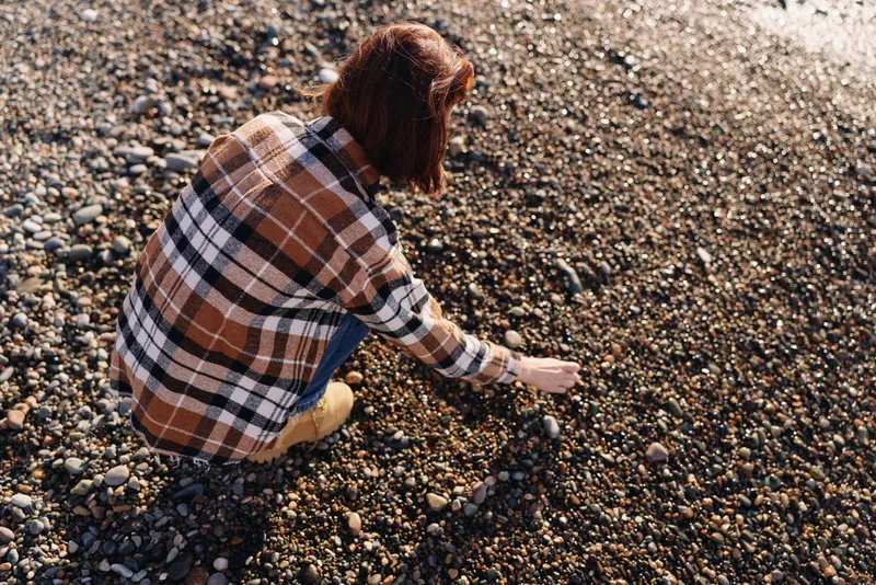 Person crouching on a pebble beach, wearing a plaid shirt, picking small stones as sunlight glints on the rocky surface in a calm outdoor scene with scattered gravel along the shore.