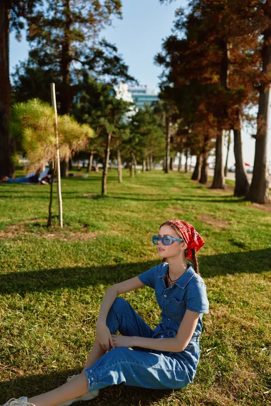 Lifestyle woman in modern boho-western denim street style with film color effect sitting outdoors in sunny park, wearing sunglasses and red bandana, relaxed mood.