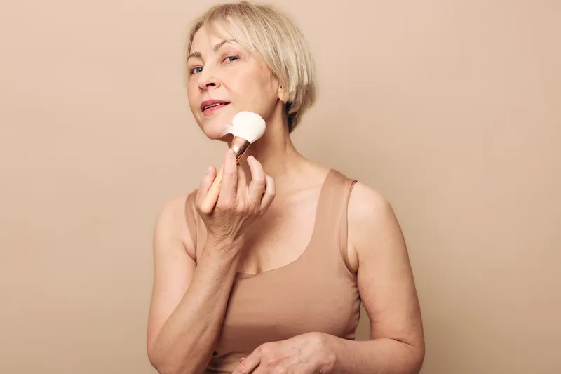 Mature woman with short blonde hair applies powder brush to face against beige background. Concept of skincare, beauty routine, self-care, aging gracefully, and natural makeup for older women.