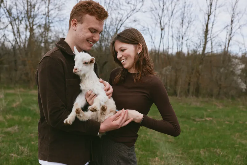 A couple holds a small goat kid in a rural field, smiling warmly as they admire their new companion. Natural light emphasizes a wholesome lifestyle moment, showcasing gentleness, care, and a strong