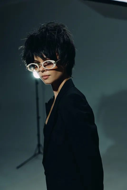 Stylish woman with short black hair wearing sunglasses and a black outfit posing against a dark studio background.