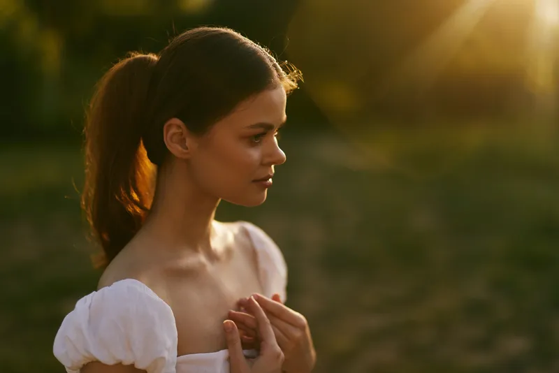 Young woman with ponytail in white dress, softly lit by golden sunlight during a serene outdoor moment, expressing contemplation and natural beauty