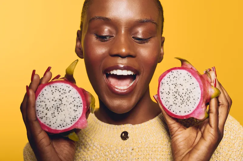 Happy dark skinned African woman holds two halves of dragon fruit, smiling with open mouth on yellow background. Concept of exotic fruits, healthy lifestyle, and natural beauty.