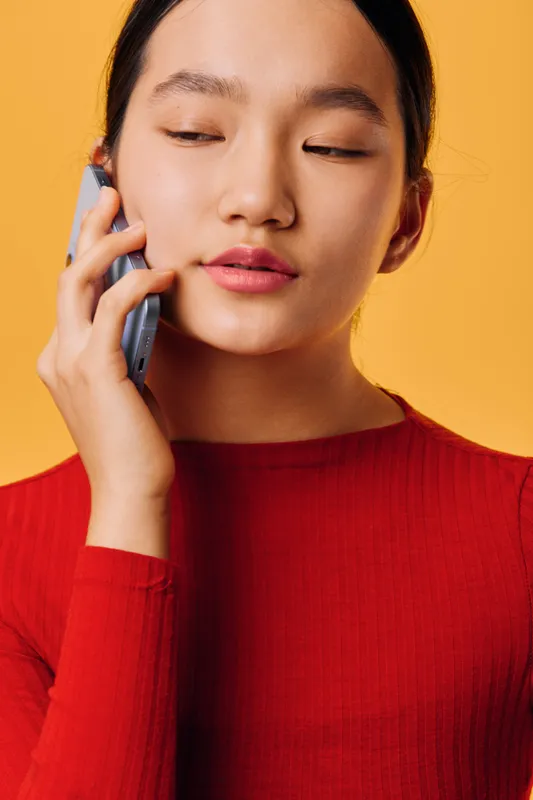 Young woman wearing red sweater talking on smartphone with eyes closed against yellow background, expressing calm and focus during mobile phone conversation in casual style.