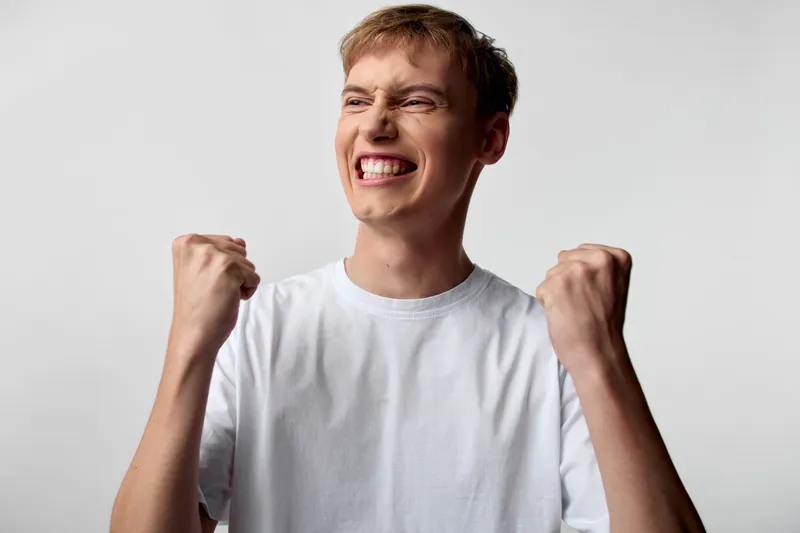 Energetic man in a white tee pumps fists in triumph, his tense shoulders and wide smile signaling victory, motivation and celebration after hard won success