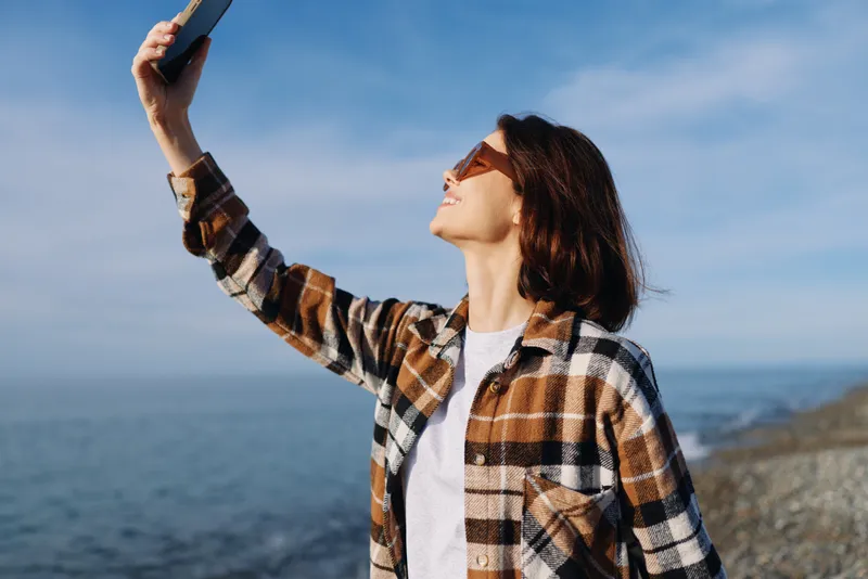 Woman outdoors coastal, holding a smartphone up, sunglasses on, plaid shirt, looking toward the sky as waves roll behind her, a bright travel and tech moment by the sea.