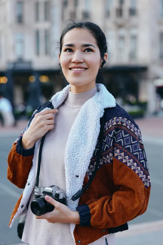 Woman with camera in an urban street scene, smiling while capturing street photography, wearing a cozy jacket, showcasing outdoor city life and everyday moments