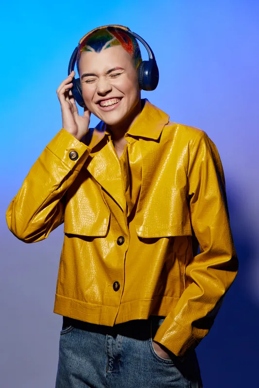 Young woman with colorful hair wearing headphones and a bright yellow jacket, enjoying music with a joyful expression on a vibrant blue and purple gradient background.