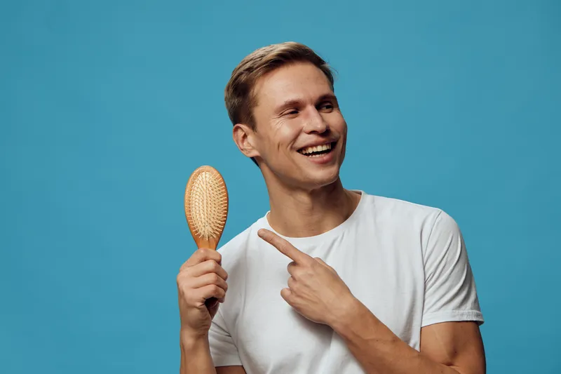 Smiling young caucasian man pointing at hairbrush wear white t-shirt isolated on blue background studio portrait mental health gadgets concept