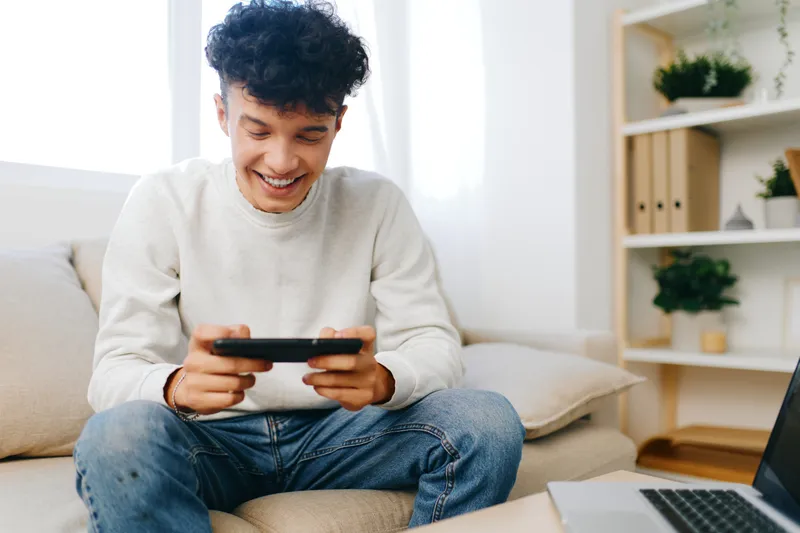 Молодой человек наслаждается игрой на смартфоне, сидя на уютном диване в современной комнате с яркими светлыми цветами и декоративными растениями