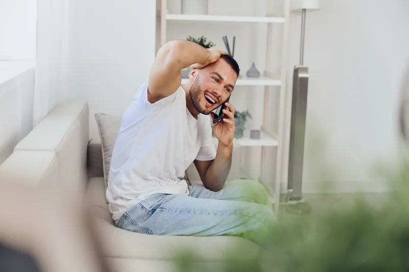 Happy man talking on the phone, sitting on a cozy couch in a modern living room, showcasing relaxation and communication. A bright and inviting atmosphere.
