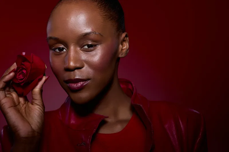 Close-up of a confident dark skinned black woman holding a red rose on a red background. Concept of beauty, elegance, romance, and natural skin glow in studio lighting.
