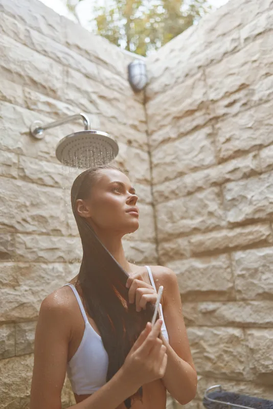 Woman showering outdoors under rain showerhead near beige stone walls, enjoying natural light and calm atmosphere with wet hair and white swimsuit top.