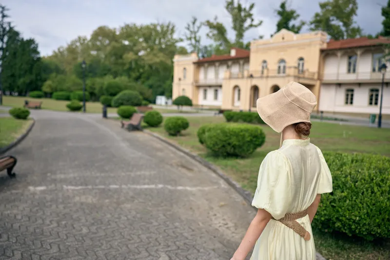 Woman wearing a vintage hat walks along a cobblestone pathway toward an elegant estate, surrounded by manicured gardens and lush outdoors scenery, capturing timeless travel vibe