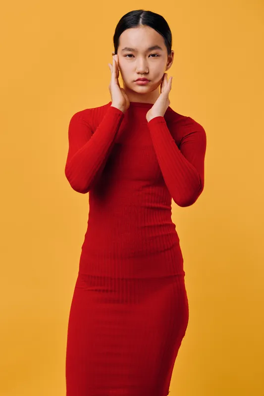 Young woman in red dress stands with hands near face against orange background, looking serious and calm with minimalist fashion style in studio portrait photography.