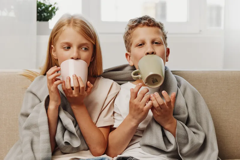 Two children wrapped in blanket drink warm beverages while sitting on couch near bright window at home.