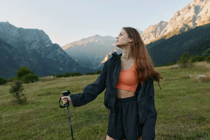 A stylish outdoor woman stands in a mountain valley with a trekking pole, gazing into the distance. She wears a bright orange top and a dark jacket, enjoying a peaceful nature moment.