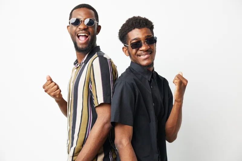 Two cheerful African American men wearing sunglasses and stylish shirts pose in a dynamic way, showcasing their playful friendship and vibrant personality.
