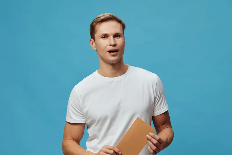 young caucasian man holding book in hand wear white t-shirt isolated on blue background looking serious thoughtful focused expression studio portrait. Education knowledge concept