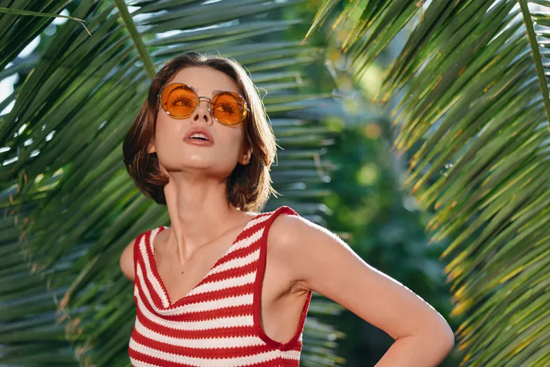 A stylish woman wearing a striped red and white sleeveless top and orange sunglasses poses outdoors among lush tropical plants, radiating confidence, vitality, and a carefree sunny mood.