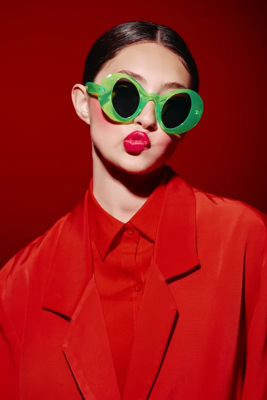 Young woman styled in bold bright red outfit with vivid green sunglasses and intense red lipstick against seamless red background. Concept for fashion, beauty, and creativity.