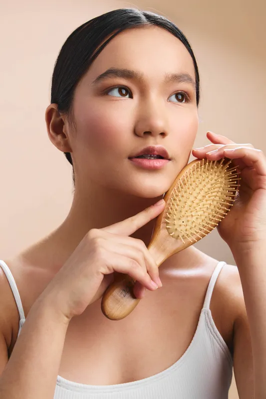 Asian woman holds a wooden hairbrush and gently grooms her hair in a softly lit space, calm and focused, creating a serene personal care moment.