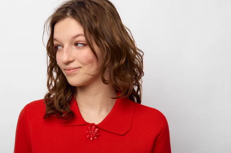 Young woman with curly brown hair wearing a red sweater looks sideways with a slight smile. Studio portrait on white background with natural expression and casual style.