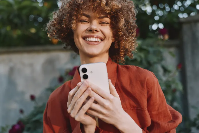 Bright portrait of a smiling woman outdoors, lifestyle moment with a phone, casual fashion and natural light, candid and joyful, greenery in the background today