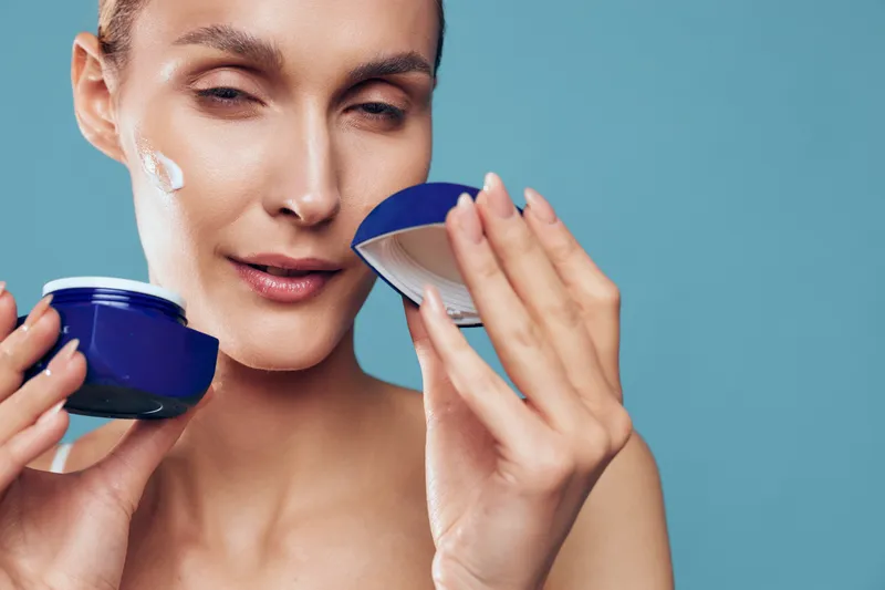 A focused woman applies facial cream in a skincare routine, applying moisturizer from a blue jar. Closeup of beauty ritual, self care and hydration in a modern setting.