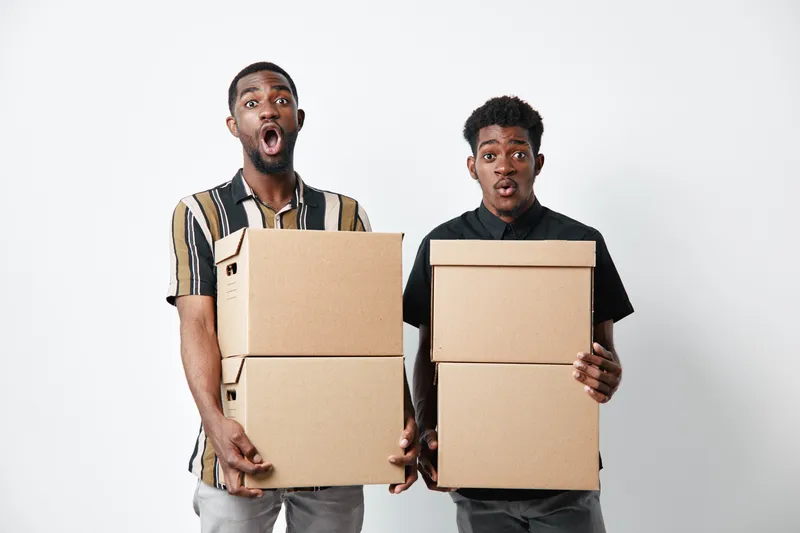 Two young black men with dark skin holding cardboard boxes with surprised expressions on white background. Casual clothing and moving or delivery concept.