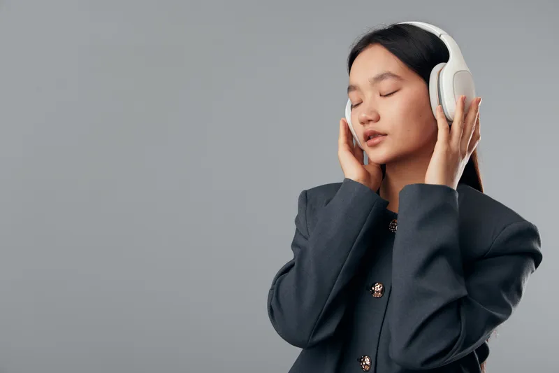 Young woman wearing headphones with closed eyes enjoying music, standing against plain background. Female portrait with long hair and relaxed expression, listening to audio content.