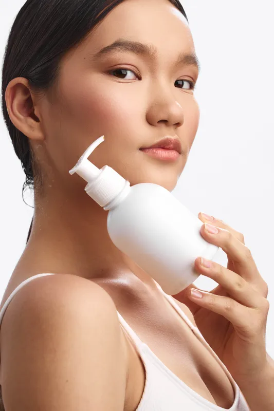 asian woman applying skincare product from a pump bottle in a closeup studio portrait. She displays clear skin and a calm expression, conveying a fresh and confident beauty routine.