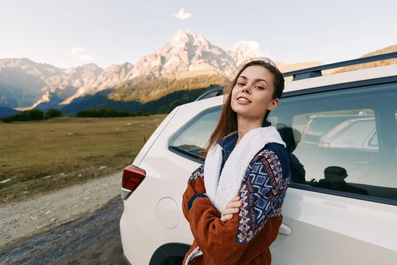 Woman, car, mountains, outdoors, travel, portrait, road, SUV. Young woman leans against a white SUV by a mountain road, wearing a cozy patterned jacket, smiling with scenic peaks and open landscape.