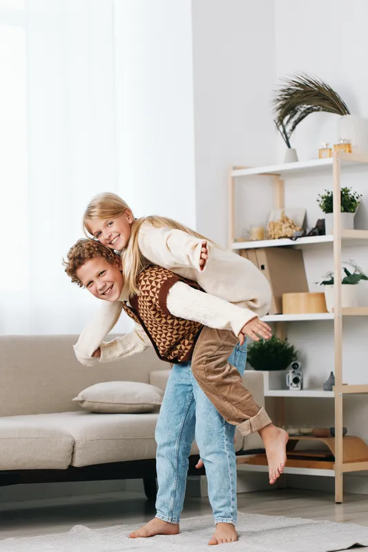 A playful couple shares a lighthearted moment indoors as they laugh and ride on each other's backs in a bright, cozy living room.