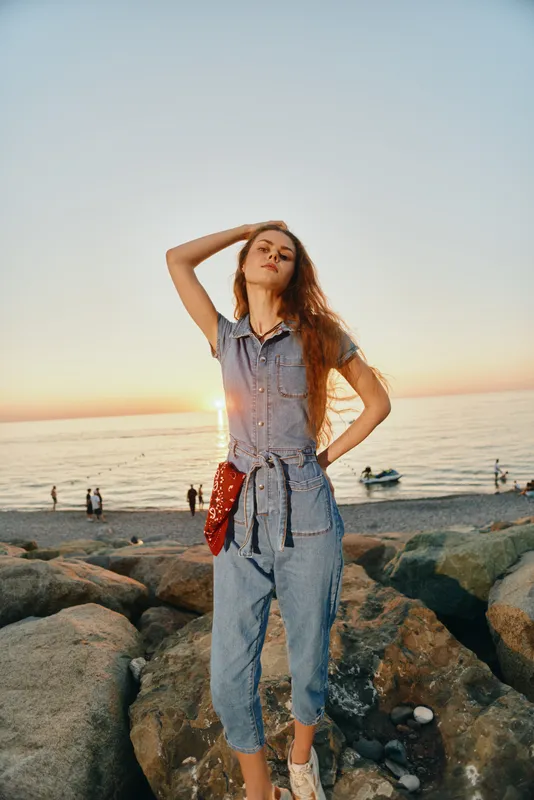 Lifestyle woman in modern boho-western denim street style posing on rocky beach at sunset with film color effect, relaxed confident mood, casual summer fashion.