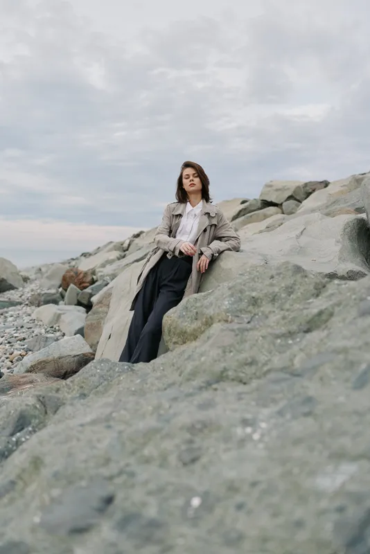 Young woman in casual outfit leans on large rocks by the sea under a cloudy sky. Natural landscape with rocky shore creates calm and serene mood in outdoor setting.