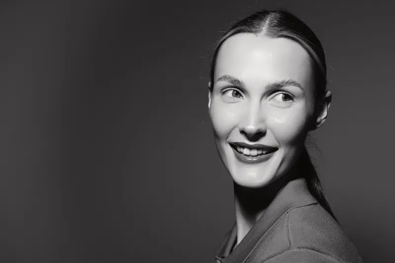 Portrait of a smiling young Caucasian woman with light skin and tied back hair looking sideways in a studio with soft lighting.