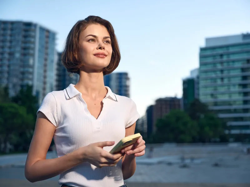 A confident business professional stands outdoors in an urban setting, texting on a smartphone, smiling and engaged, projecting a positive, dynamic mood and modern workplace energy.