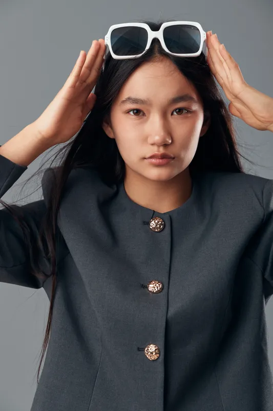 Young woman with long black hair wearing dark jacket and white sunglasses on head posing in studio. Portrait, fashion and beauty concept with serious expression against gray background.