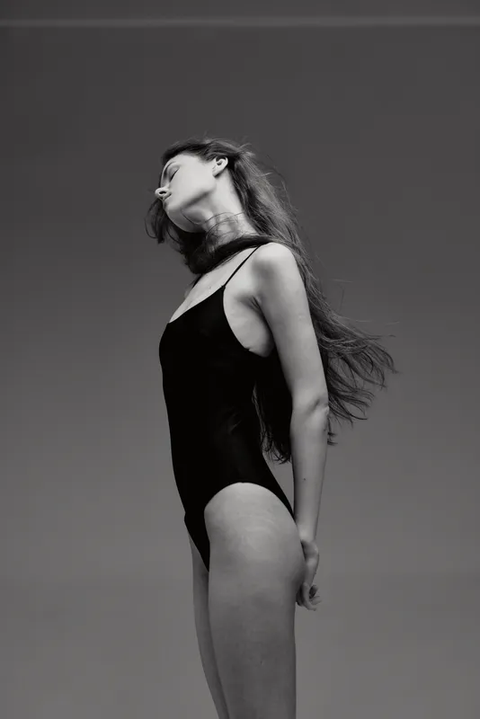 Black and white artistic portrait of a woman in a swimsuit posing with closed eyes and flowing hair against clear sky.