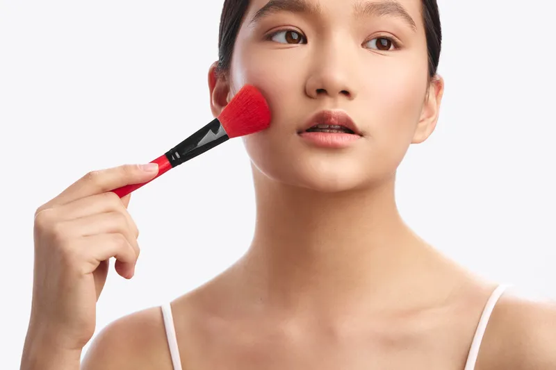 asian woman applies makeup with a brush in a clean studio, focusing on her complexion. she smiles softly, conveying a calm, confident mood and a moment of personal care.