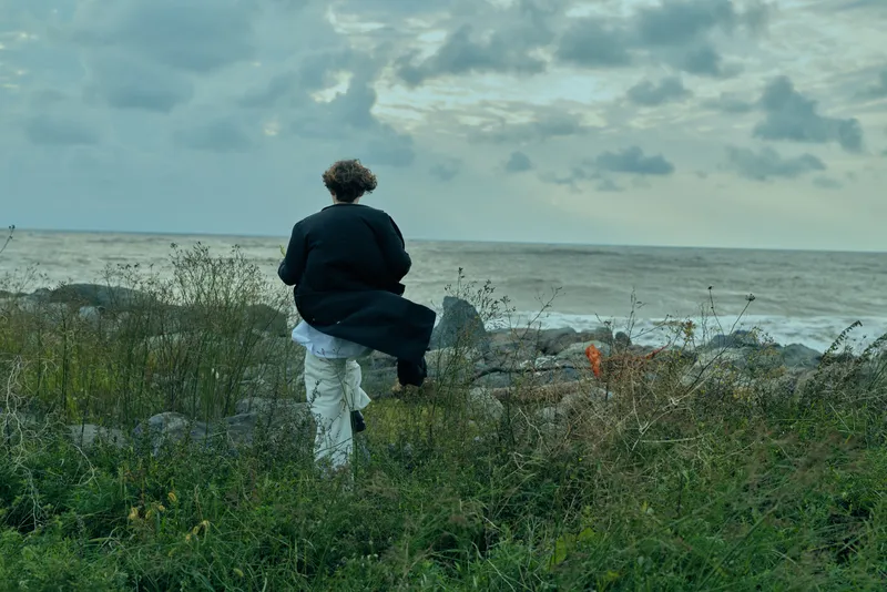 Walking figure in a dark coat along a windswept coastal meadow toward the sea under a cloudy sky, emphasizing solitude, exploration, quiet resilience, and the expansive horizon.