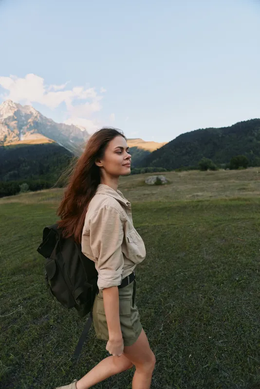 A young woman with a backpack walks through a grassy field near mountains under a clear sky. Concept of outdoor lifestyle, hiking, and nature exploration.