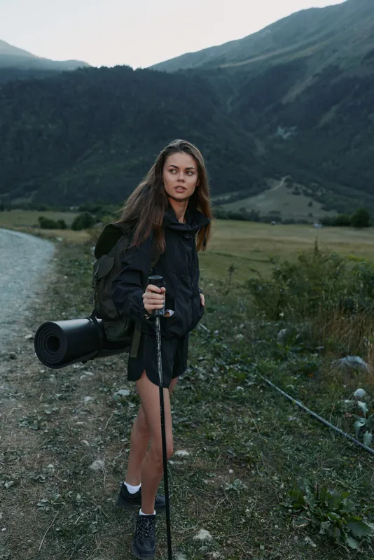A female hiker stands on a rocky trail beside a mountain stream, holding a camera and trekking pole. She wears a dark jacket and shorts, ready to explore rugged scenery with a calm, focused demeanor.