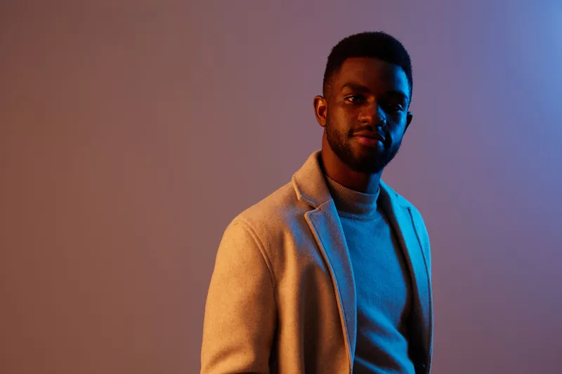 Portrait of a confident young Black man wearing a stylish sweater and blazer against a vibrant background with soft blue and orange lighting, embodying modern fashion
